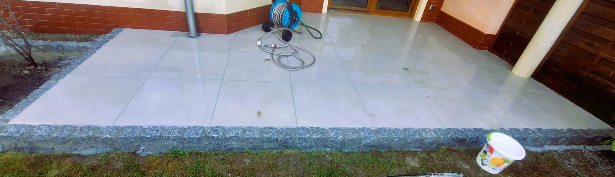 Wykończony taras z jasnych płyt, obrzeża z kostki granitowej. Widoczny fragment elewacji budynku i narzędzia pracy. Całość w jasnej tonacji, z lekkim połyskiem.