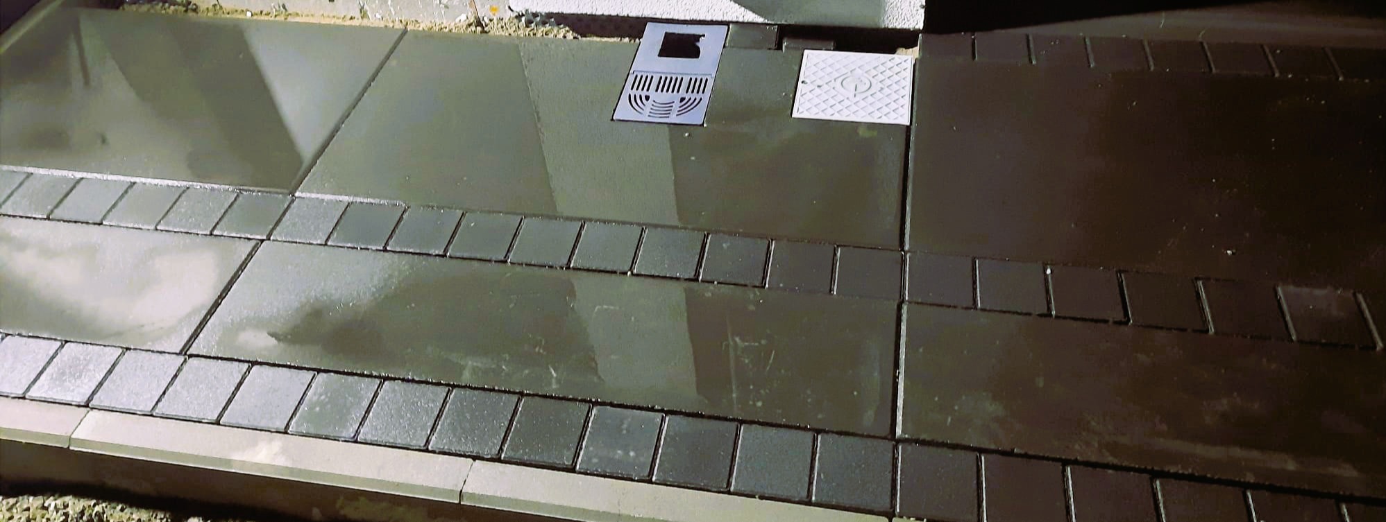 Świeżo ułożona, ciemna kostka brukowa z odpływem liniowym i studzienką rewizyjną. Widoczne obrzeża i fuga. Całość sprawia wrażenie mokrej, być może po myciu.