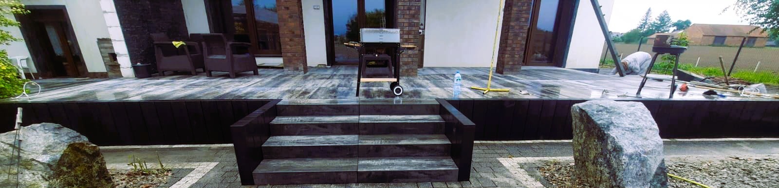 Nowoczesny taras z szarej deski kompozytowej, z niskimi schodami i dużymi kamieniami dekoracyjnymi. W tle budynek z dużymi oknami i sprzęt do grillowania.