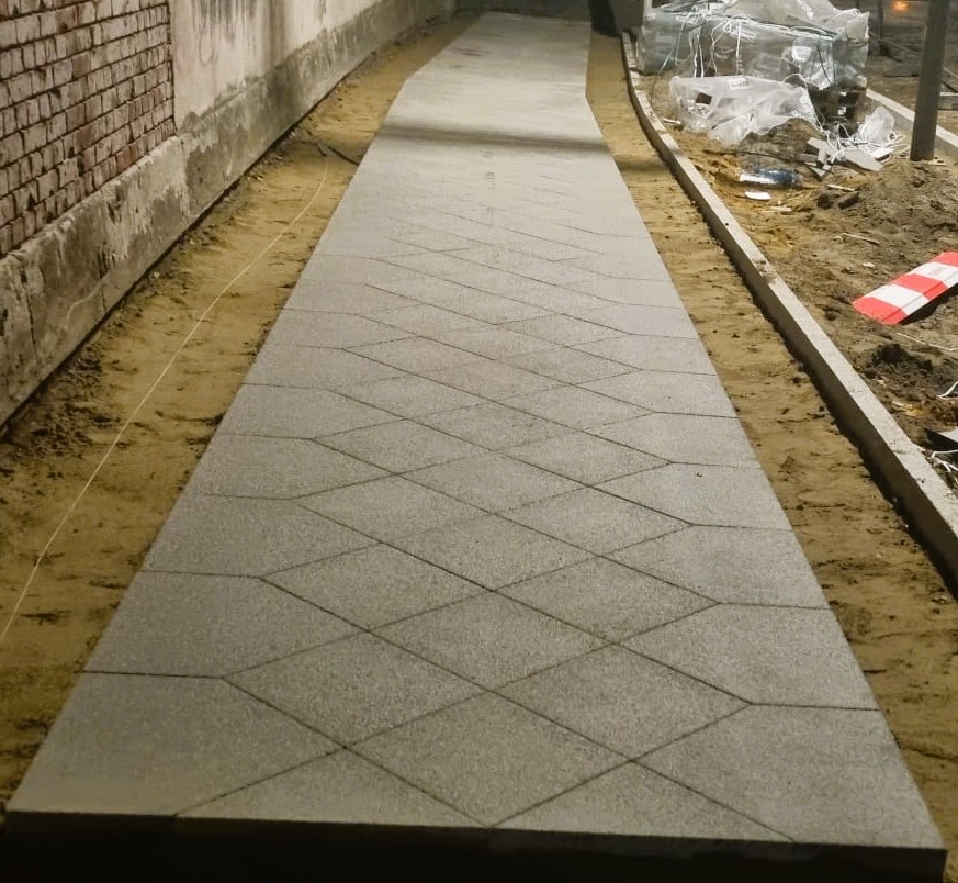 Ścieżka z szarej kostki o wzorze rombów, otoczona piaskiem i ceglanym murem. Widoczne materiały budowlane w tle. Ujęcie z perspektywy.