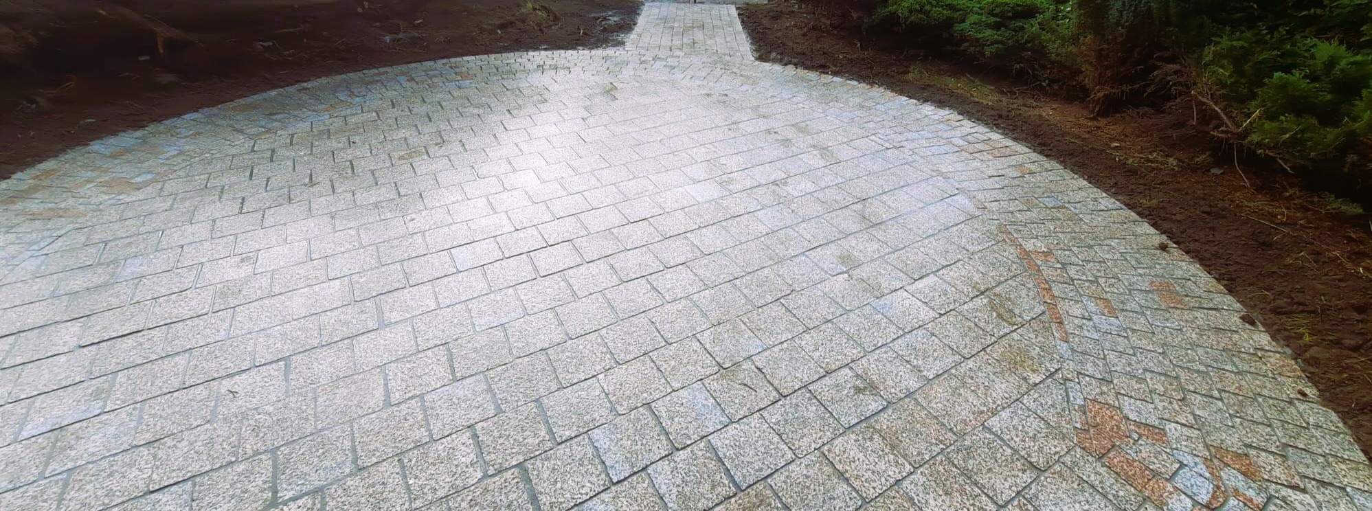 Ścieżka z jasnoszarej kostki granitowej ułożona łukiem, otoczona ziemią i zielenią. Widoczny fragment ścieżki z kostki w odcieniach brązu.