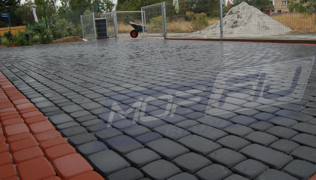 Ułożona, mokra, grafitowa kostka brukowa z bordurą z czerwonej kostki. W tle ogrodzenie, wózek i pryzma piasku. Widok z perspektywy.