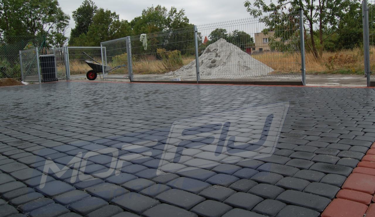 Ułożona, mokra kostka brukowa w odcieniach szarości z czerwoną obwódką. W tle ogrodzenie z siatki, taczka i pryzma piasku. Pochmurny dzień.