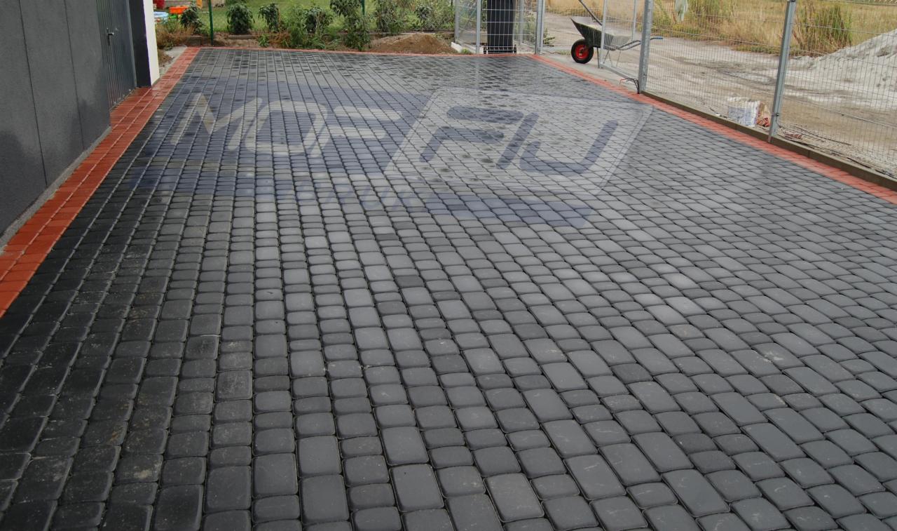 Ułożona, szara kostka brukowa z czerwoną obwódką przy garażu. W tle widoczny wózek budowlany i piasek. Całość tworzy estetyczne wykończenie podjazdu.