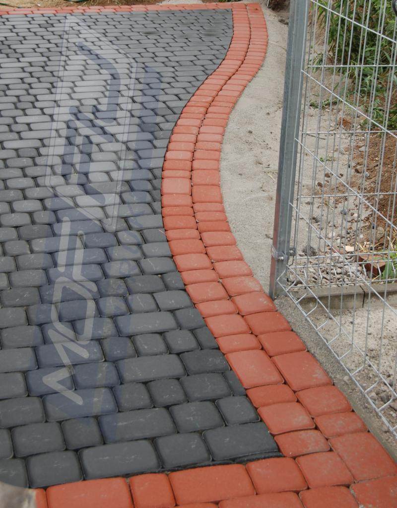 Ścieżka z kostki brukowej w kolorze szarym, obramowana czerwoną kostką, biegnąca wzdłuż metalowej siatki ogrodzeniowej. Widok z góry, z bliska.