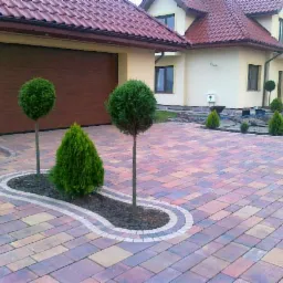 Kolorowa kostka brukowa przed domem z garażem, ozdobne nasadzenia drzew i krzewów w wydzielonej rabacie. Widok na budynek z czerwonym dachem.