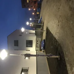 Nowe lampy oświetleniowe wzdłuż chodnika przed budynkiem mieszkalnym, częściowo owinięte ochronną folią, sfotografowane o zmierzchu.