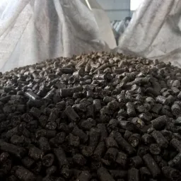 Góra ciemnego, brązowo-czarnego pelletu drzewnego w worku jutowym. Widok z bliska na strukturę i kształt granulatu. Oświetlenie magazynowe.