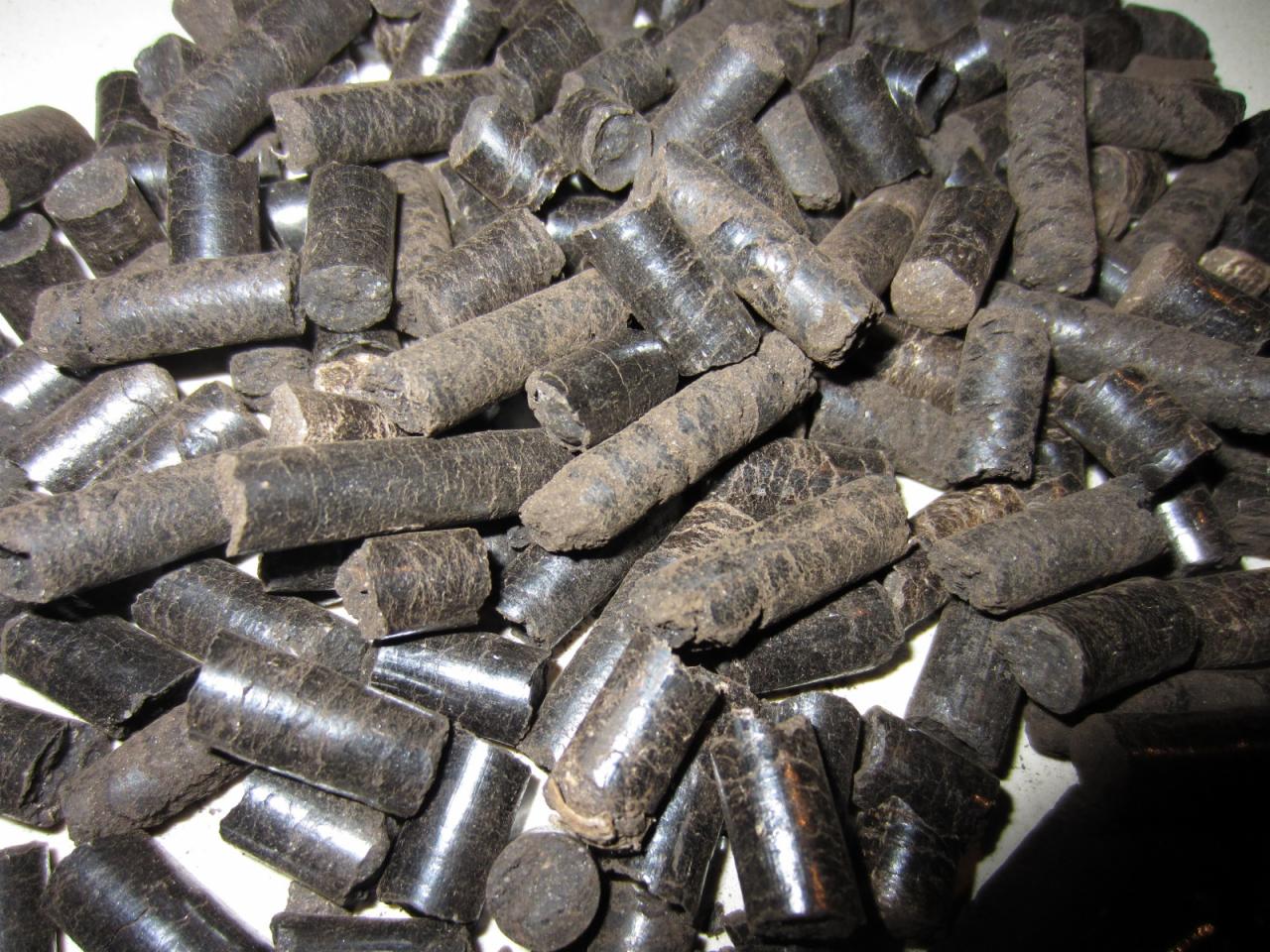 Kupa ciemnych, cylindrycznych bryłek pelletu torfowego 6 mm, z widoczną strukturą włókien i nierówną powierzchnią, ułożona na jasnym podłożu, oświetlona z boku.