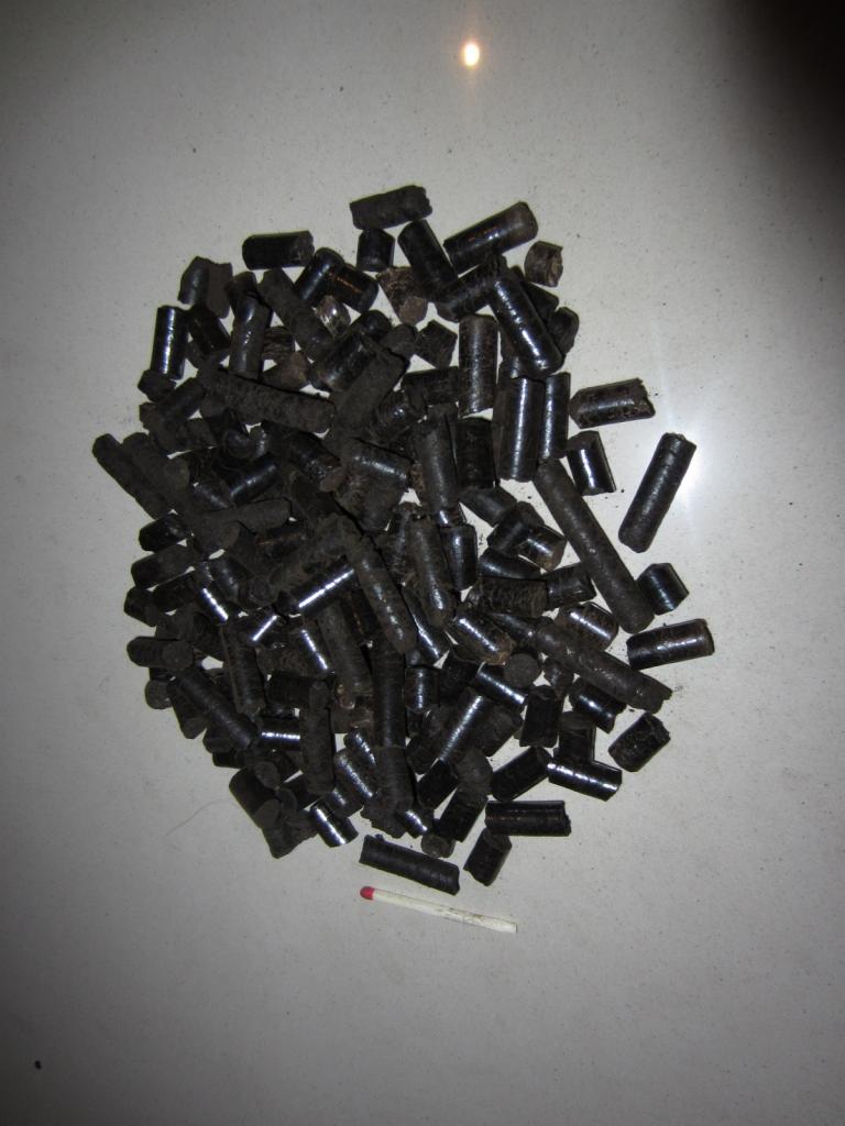 Kupka ciemnego pelletu torfowego o średnicy 6 mm na szarej powierzchni, obok leży zapałka dla porównania skali.
