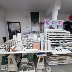 Ekspozycja różnorodnych płytek ceramicznych w sklepie, ułożonych na stole i regałach, z grafiką przedstawiającą rzeźby z gumami balonowymi w tle.