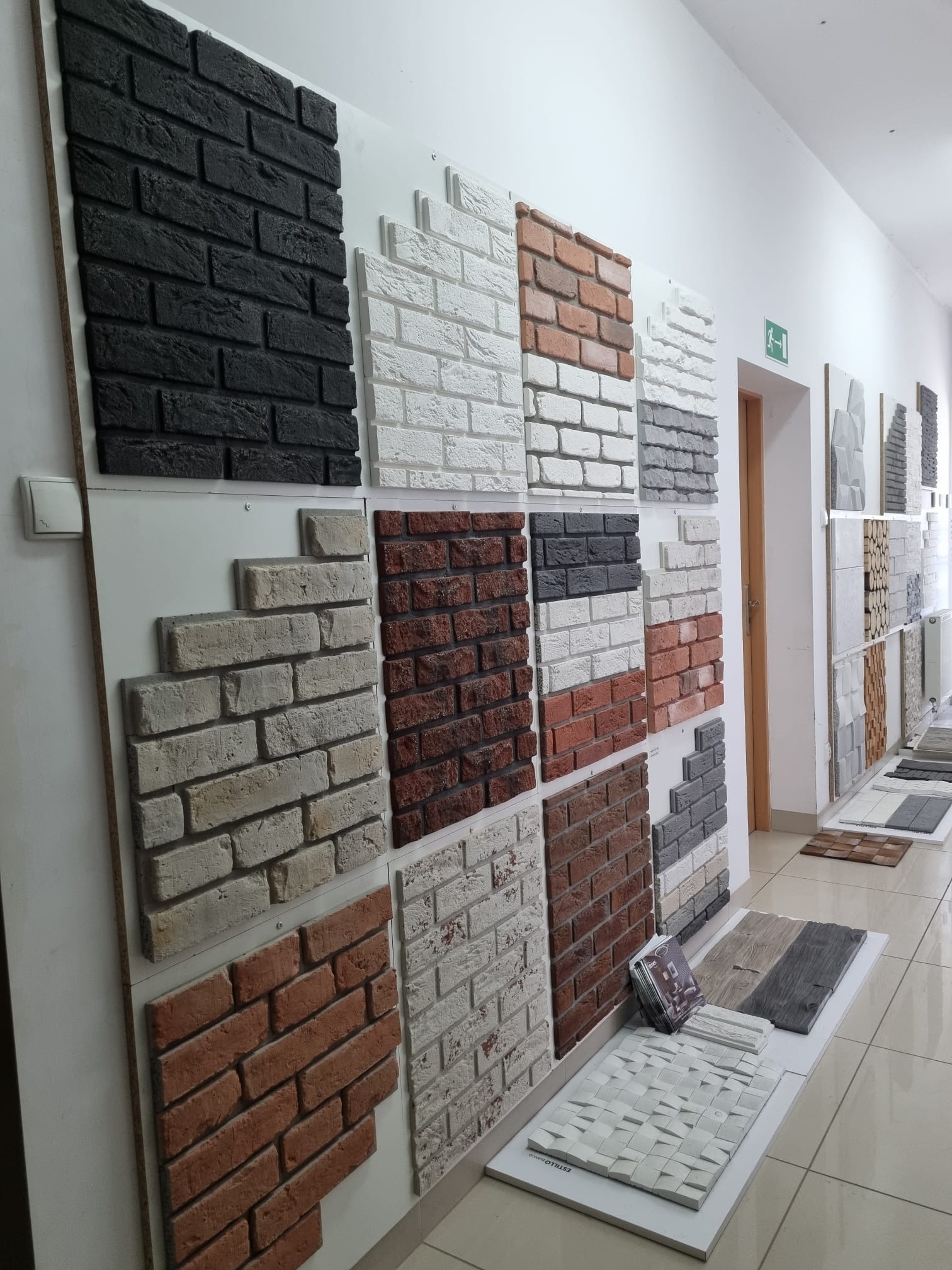 Ekspozycja różnorodnych paneli dekoracyjnych imitujących cegłę w odcieniach czerni, bieli, szarości i czerwieni, prezentowanych na białych płytach w salonie z płytkami.