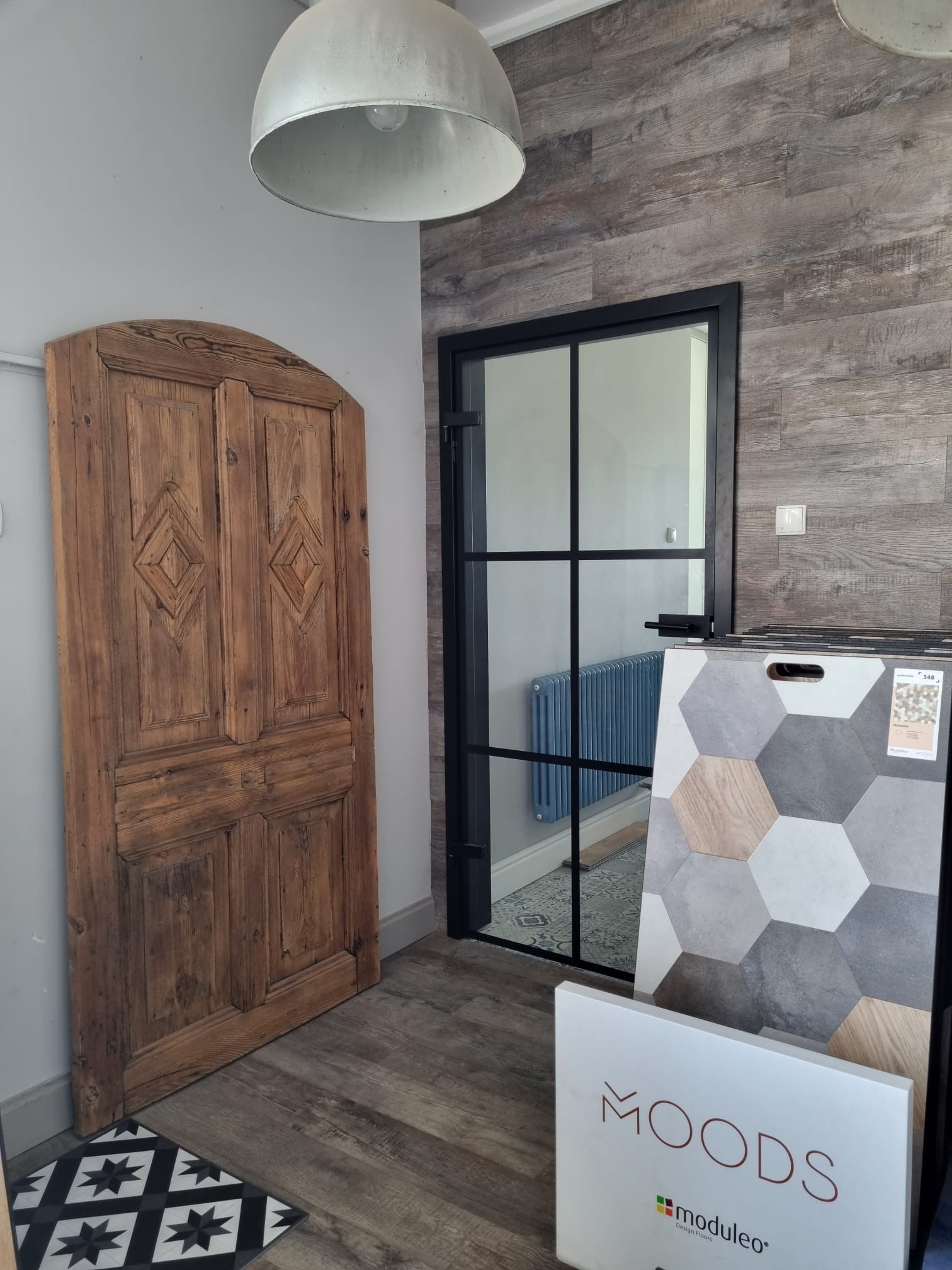 Wnętrze prezentujące starą drewnianą stolarkę drzwiową, panele ścienne imitujące drewno, wzornik płytek heksagonalnych oraz grzejnik za przeszklonymi drzwiami w czarnej ramie.