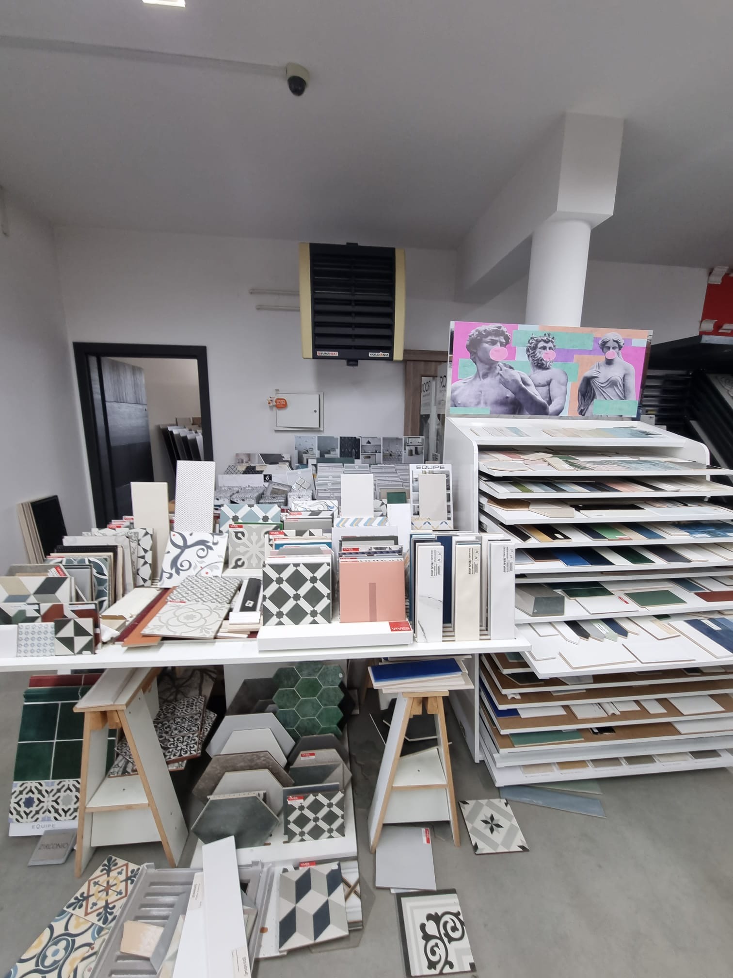 Ekspozycja różnorodnych płytek ceramicznych w sklepie, ułożonych na stole i regałach, z grafiką przedstawiającą rzeźby z gumami balonowymi w tle.