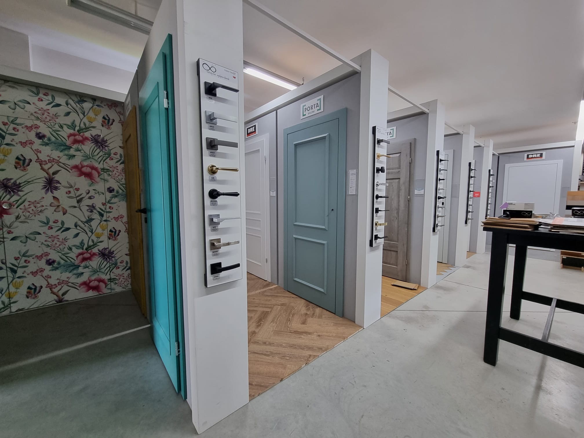 Ekspozycja różnych modeli drzwi wewnętrznych w salonie, z panelami prezentującymi klamki i okucia, widoczne drzwi w kolorze turkusowym i szarym.