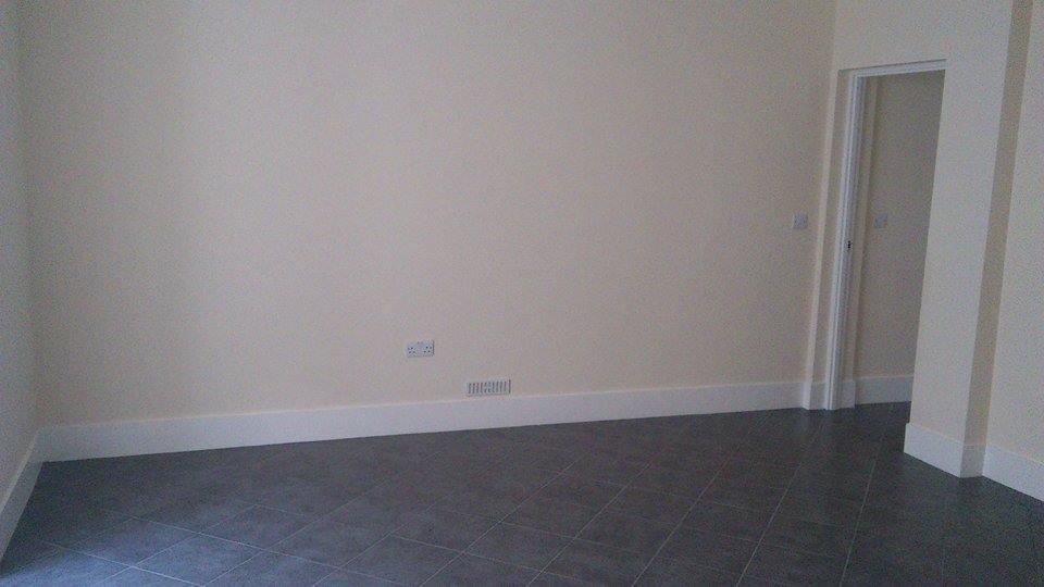 Pomalowane na kremowo ściany, szara podłoga z płytek i białe listwy przypodłogowe w pustym pokoju z otwartymi drzwiami w tle.