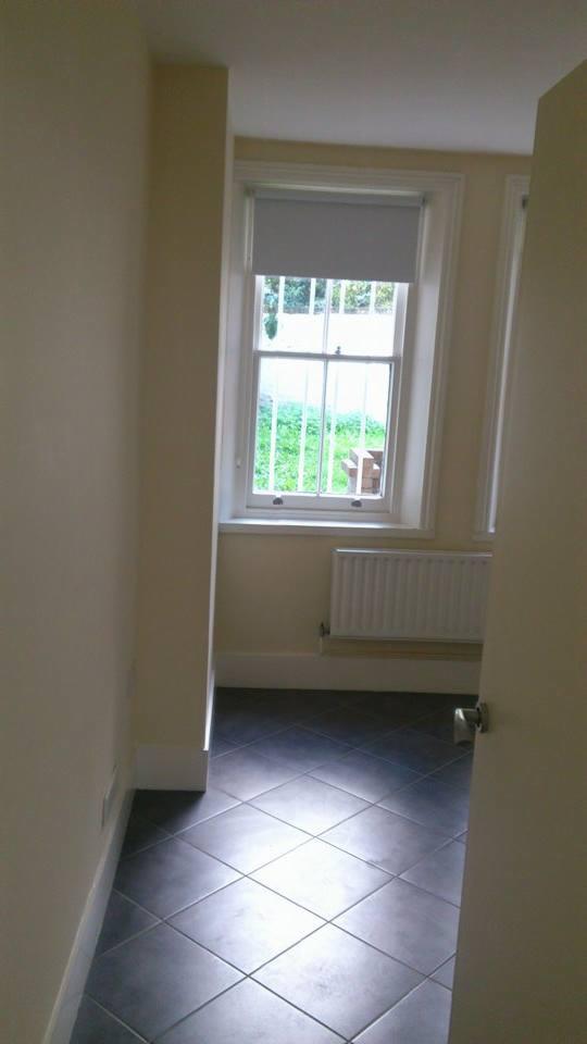 Wnętrze pomieszczenia z ciemną, błyszczącą podłogą ułożoną w karo, jasnymi ścianami i oknem z roletą.