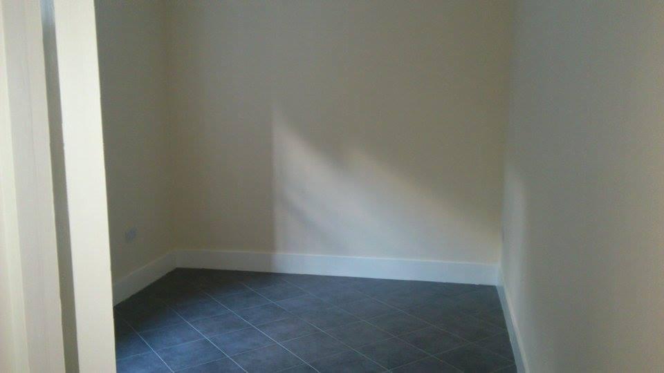 Wnętrze pomieszczenia z ciemną, kwadratową podłogą z płytek, białymi listwami przypodłogowymi i jasnymi ścianami, widok zza białej framugi drzwi.