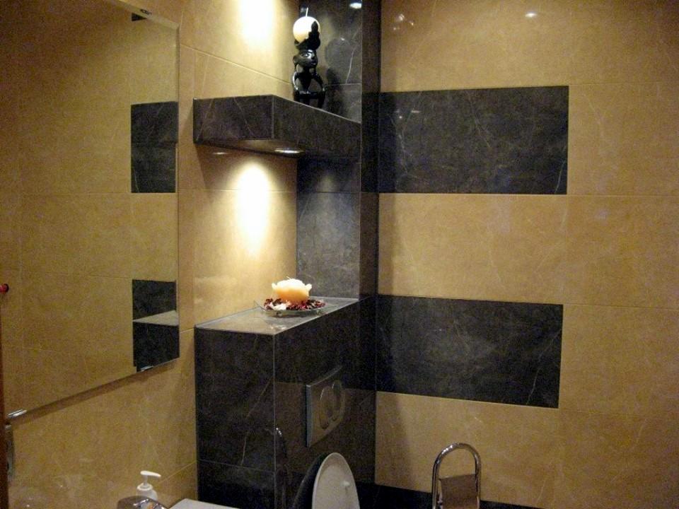 Ukończona łazienka z beżowo-czarnymi płytkami, podświetlana półka z dekoracją, widoczna umywalka i toaleta.