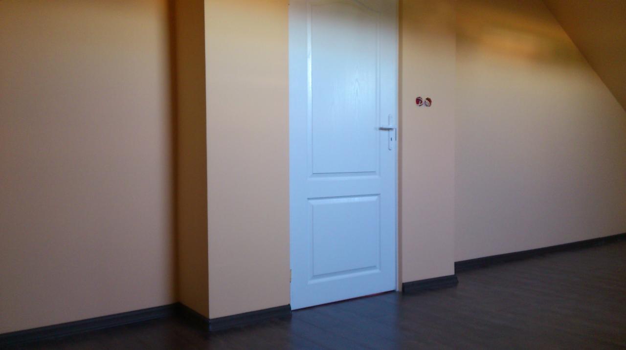 Wnętrze pokoju na poddaszu z pomalowanymi na beżowo ścianami, ciemną podłogą i zamontowanymi białymi drzwiami z srebrną klamką, widoczne gniazdka elektryczne bez osprzętu.