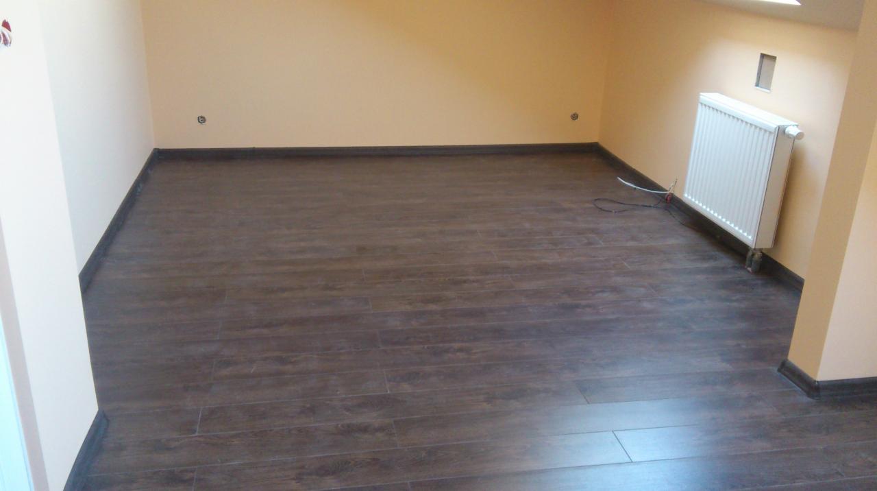 Pokój po remoncie z panelami podłogowymi imitującymi drewno, kremowe ściany z widocznymi gniazdkami i biały grzejnik pod oknem na poddaszu.