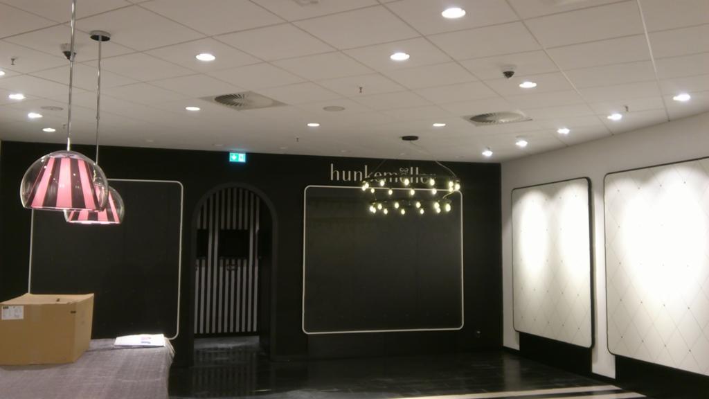 Wnętrze sklepu w trakcie prac wykończeniowych: czarne ściany z białymi obramowaniami, dekoracyjne oświetlenie, podwieszane lampy w paski, widoczny karton na podłodze.