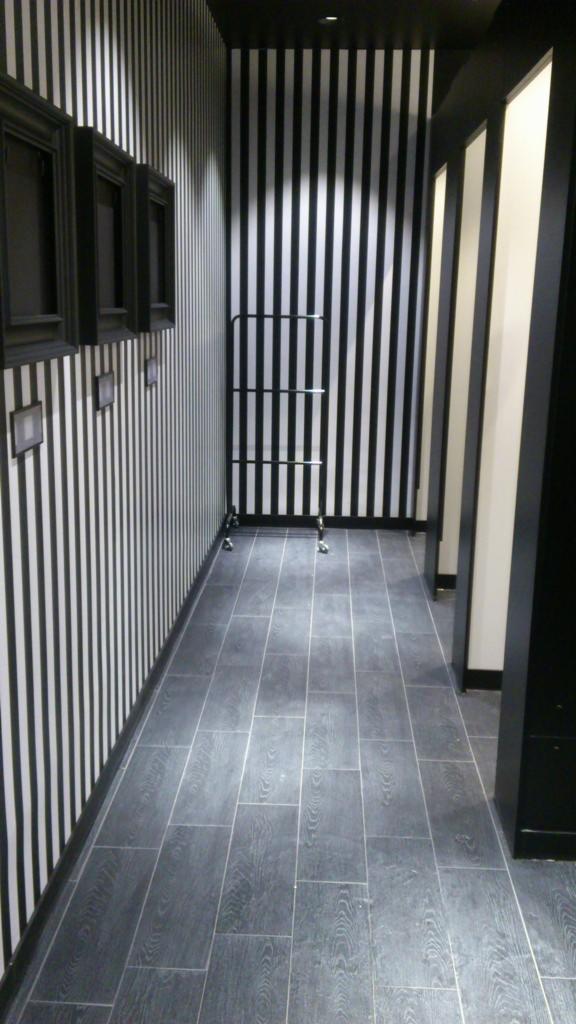 Wnętrze w czarno-białe pasy z szarą podłogą z płytek imitujących drewno, czarne ramy na ścianie i metalowy wieszak na kółkach.