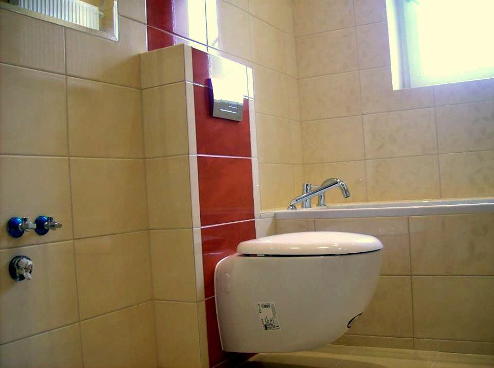 Nowoczesna łazienka z podwieszaną toaletą, beżowe płytki ceramiczne z dekoracyjnym czerwonym pasem, bateria wannowa na wannie, okno w tle.