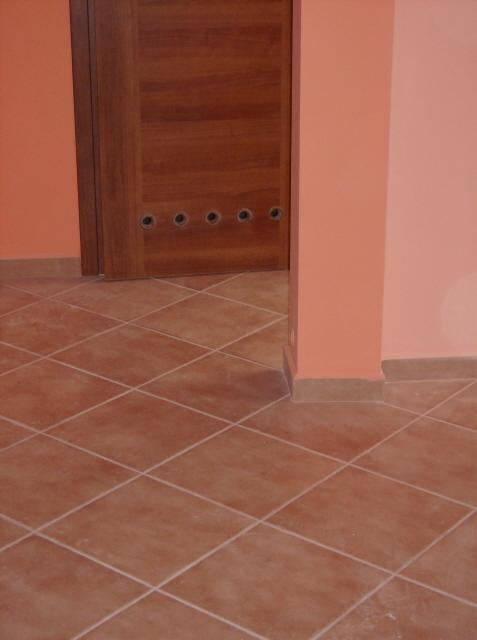 Wykończona podłoga z terakoty ułożonej w karo, widoczny fragment drewnianych drzwi z rzędem otworów wentylacyjnych w dolnej części oraz fragment ściany pomalowanej na kolor brzoskwiniowy.