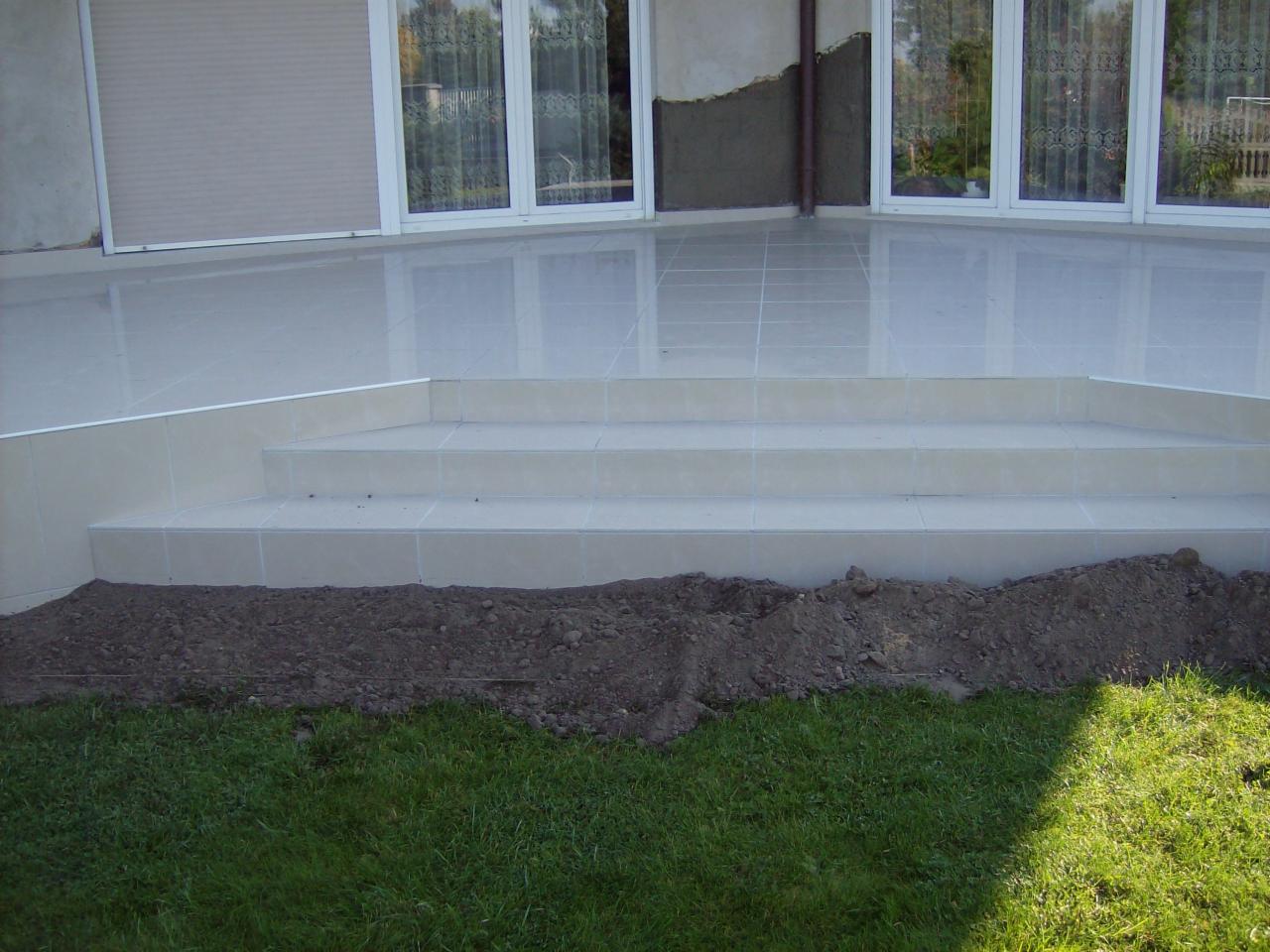 Wyłożone jasnymi płytkami schody prowadzące do tarasu, z widocznym fragmentem świeżo nasypanej ziemi i trawnika u dołu kadru, w tle okna z roletami.