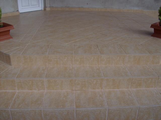 Beżowe płytki ceramiczne na schodach wejściowych i tarasie, wzór ułożony pod kątem, donice z roślinami po bokach.