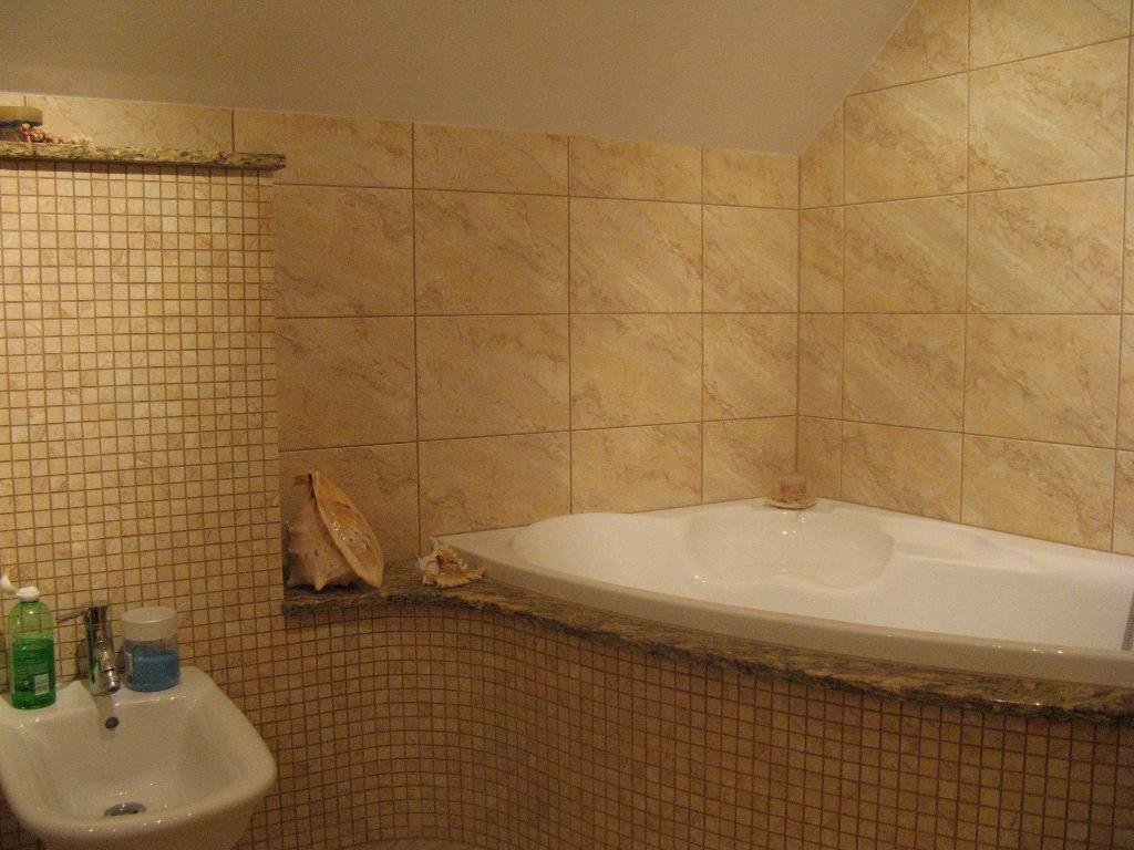 Wykończona łazienka z narożną wanną obłożoną beżowymi płytkami imitującymi marmur, mozaika na ścianie obok umywalki, dekoracyjne muszle.