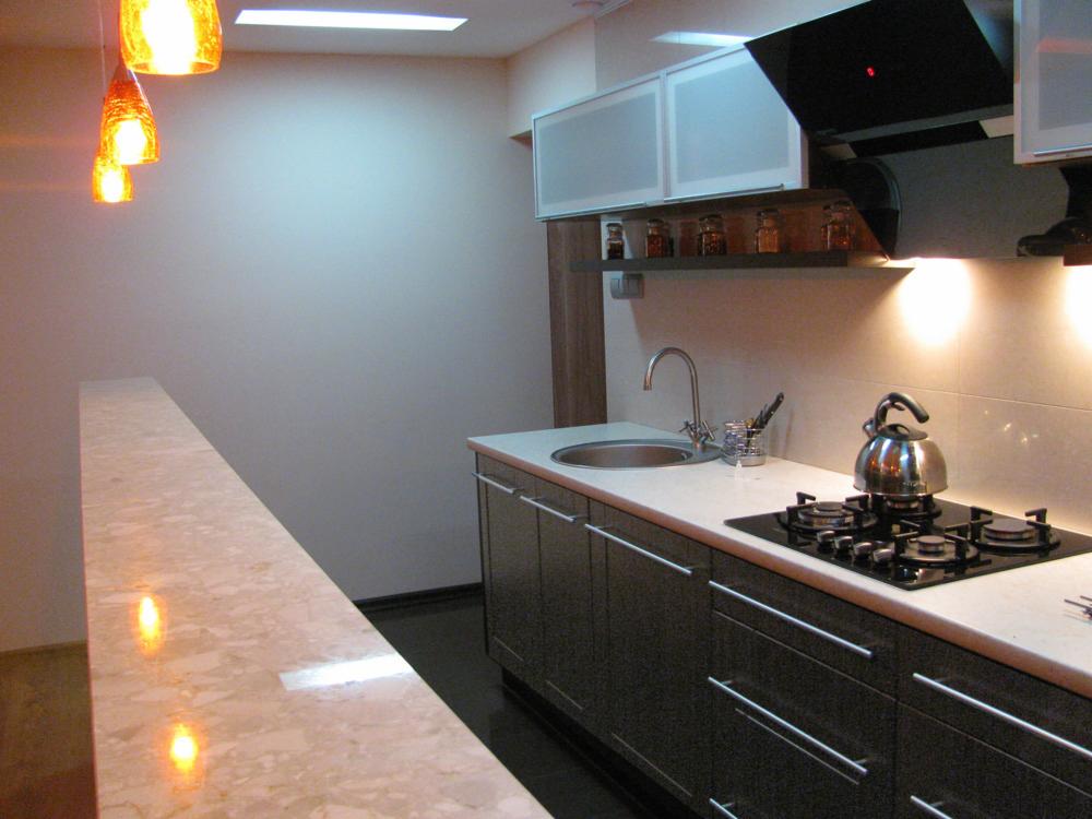 Nowoczesna kuchnia z czarnymi szafkami, jasnym blatem, okrągłym zlewem i płytą gazową z czajnikiem, oświetlona wiszącymi lampami o ciepłej barwie.