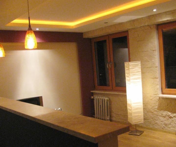 Nowoczesne wnętrze z podwieszanym sufitem z oświetleniem LED, kamienną ścianą i drewnianymi oknami, uzupełnione wiszącymi lampami i wysoką lampą podłogową z abażurem.