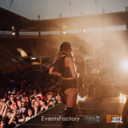 Scena na stadionie podczas koncertu plenerowego, tłum publiczności i oświetlenie sceniczne, wokalistka w czarnym stroju odwrócona tyłem do widza.