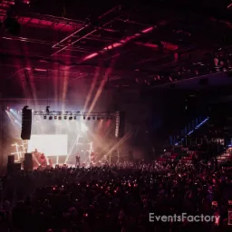 Scena koncertowa z zespołem na tle jasnego ekranu i tłumu publiczności, oświetlona reflektorami w odcieniach różu i bieli.