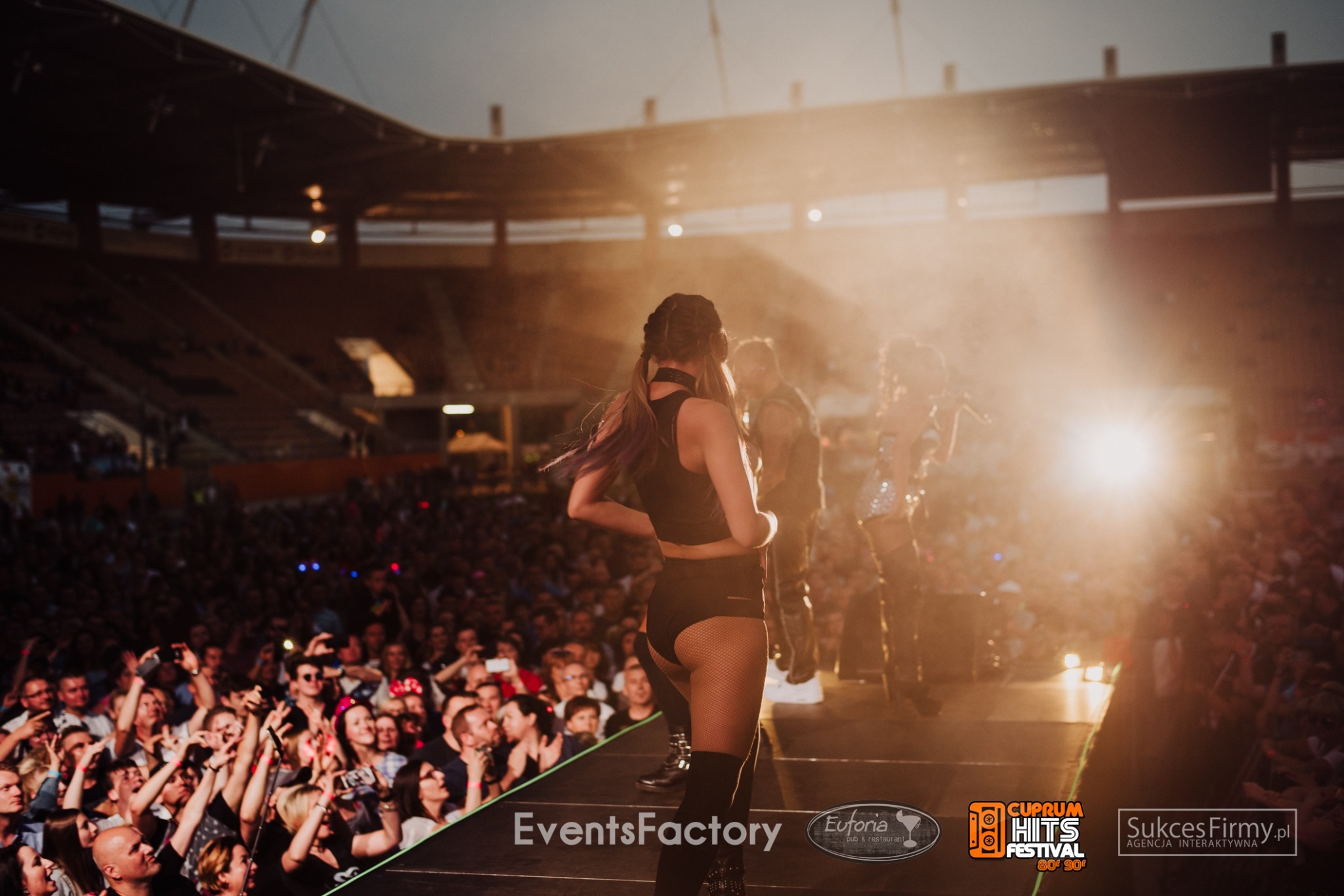 Scena na stadionie podczas koncertu plenerowego, tłum publiczności i oświetlenie sceniczne, wokalistka w czarnym stroju odwrócona tyłem do widza.