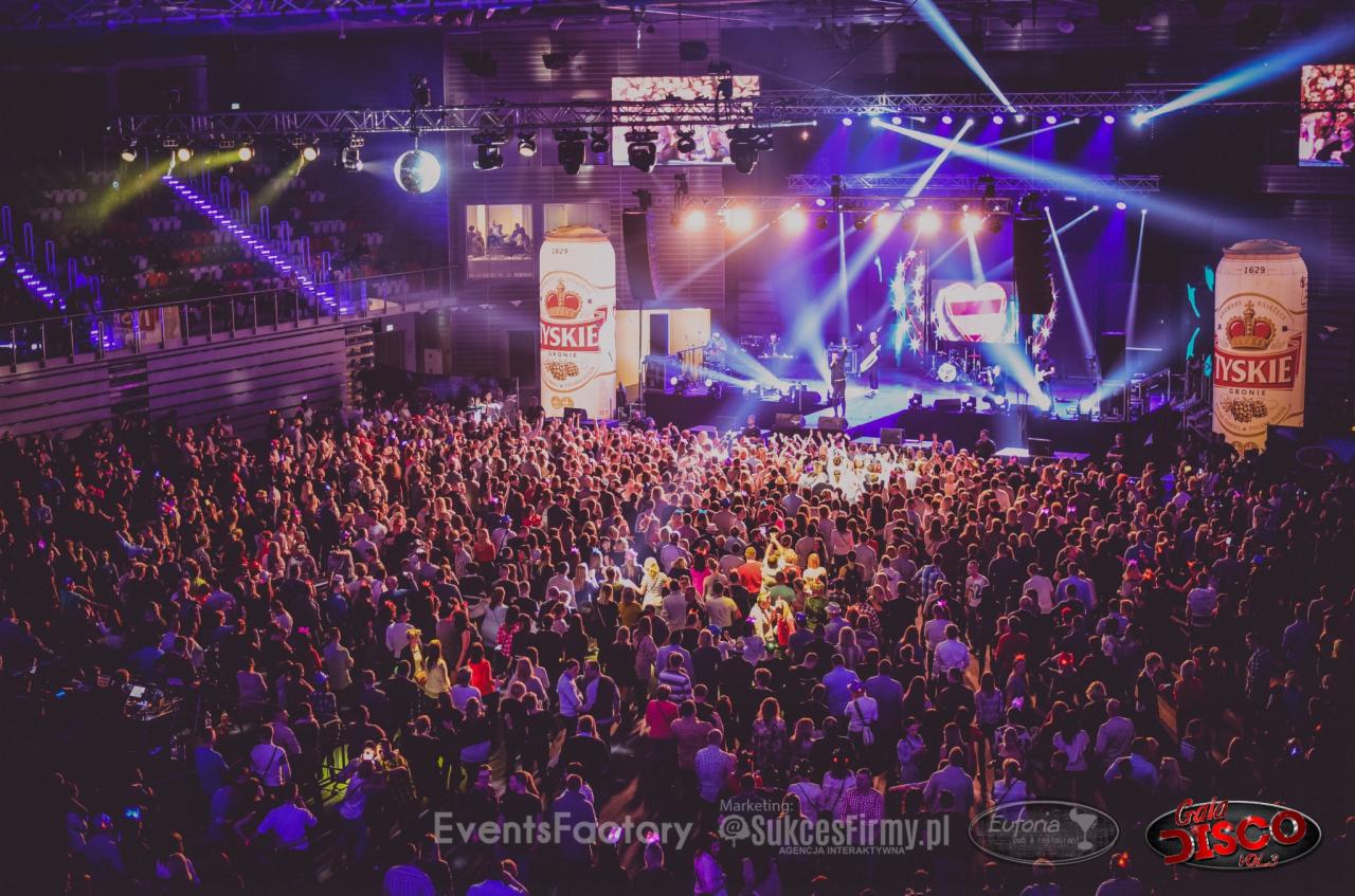 Widok z góry na duży tłum ludzi bawiących się na koncercie w hali, scena oświetlona reflektorami, gigantyczne atrapy puszek piwa po bokach sceny, lustrzana kula zawieszona pod sufitem.