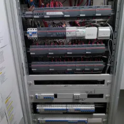 Rozdzielnica elektryczna w metalowej szafie z licznymi przewodami, zabezpieczeniami i modułami sterującymi, widoczne etykiety opisowe.