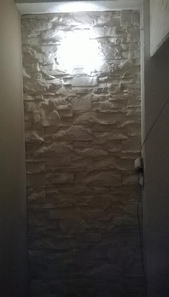 Ściana wykończona dekoracyjnym, jasnym kamieniem w wąskim przejściu, oświetlona od góry, tworząca efektowną teksturę. Widoczne gniazdko elektryczne.