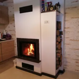 Nowoczesny, biały piec z obudową z ciemnego kamienia i wnęką na drewno, w tle ściana wyłożona kamieniem dekoracyjnym i fragment zabudowy kuchennej.