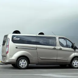 Beżowy van z logo 'TRANS PORCIKS' na tle białego budynku. Perspektywa boczna, widoczne koła i detale karoserii.