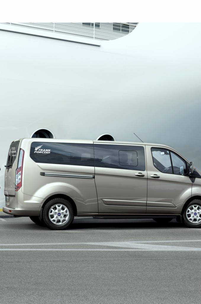 Beżowy van z logo 'TRANS PORCIKS' na tle białego budynku. Perspektywa boczna, widoczne koła i detale karoserii.