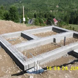 Zalane betonem fundamenty domu jednorodzinnego z wystającymi prętami zbrojeniowymi, widoczne niebieskie rury i data wykonania w prawym dolnym rogu, w tle zalesione wzgórza.