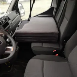 Wnętrze dostawczego samochodu z szarą, składaną leżanką/materacem zamontowanym nad siedzeniami pasażera, widok na kierownicę i deskę rozdzielczą.