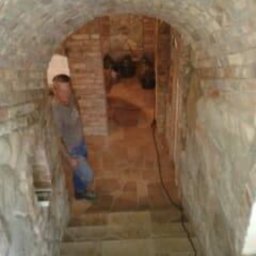 Pan - Murowany łuk z cegły, schody z kamienia, mężczyzna w kasku. Kamienna konstrukcja przypomina piwnicę lub podziemne przejście. Widoczne fugi i naturalna kolorystyka materiałów.