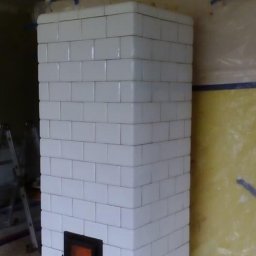 Pan - Biały piec kaflowy w trakcie budowy, widoczne zabezpieczenia ścian folią i taśmą malarską, niedokończone wykończenie sufitu.