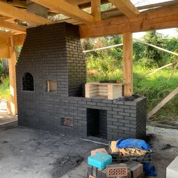 Pan - Ciemna, ceglana konstrukcja kominka zewnętrznego w trakcie budowy, z widocznymi elementami szkieletu drewnianego i narzędziami murarskimi wokół. Detal ceglanego wykończenia.