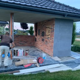 Pan - Budowa pieca chlebowego z cegły na zewnątrz. Widoczny murarz w trakcie pracy, narzędzia i materiały budowlane. Częściowo zadaszona konstrukcja z ciemną dachówką.