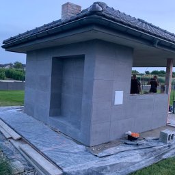 Pan - Betonowa altana z kominem w trakcie budowy, widoczne płyty elewacyjne, drewniane elementy konstrukcyjne i narzędzia. Dwóch mężczyzn w tle.